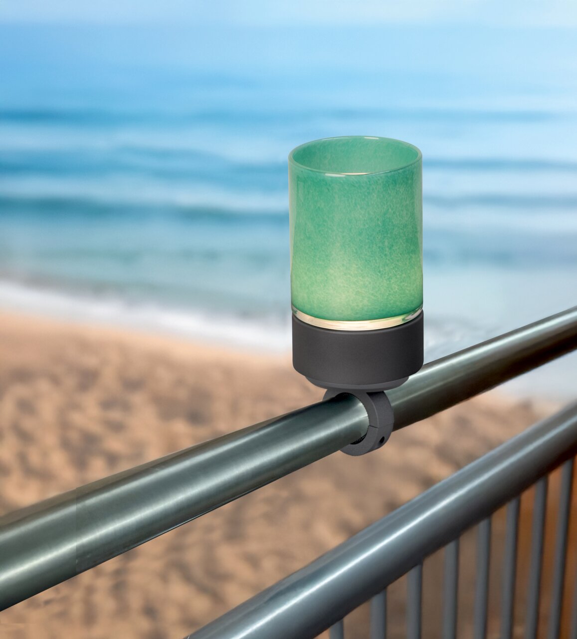 Geländerhalterung mit grünem Akku-Windlicht, montiert an einem Metallgeländer mit Blick auf das Meer im Hintergrund.