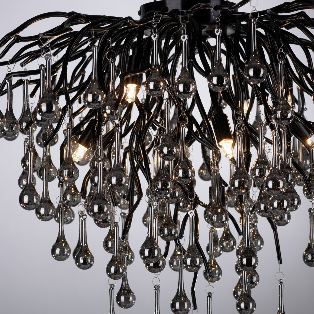 Close-up van een moderne plafondlamp met veel hangende, druppelvormige glazen elementen en brandende gloeilampen, gezien van onderaf.