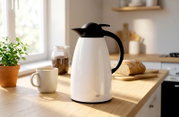 Witte Alfi isoleerkan/thermoskan met zwarte deksel en handgreep op een houten aanrecht in een lichte keuken, gefotografeerd in een driekwart vooraanzicht vanaf ooghoogte; naast een koffiekopje.