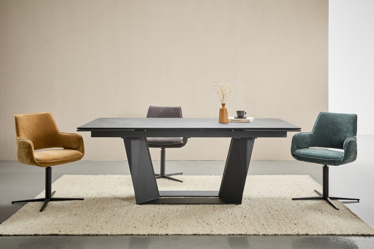 Moderne eettafel met een zwart onderstel en een grijs tafelblad, omringd door drie gekleurde stoelen in een lichte ruimte, gezien van voren.