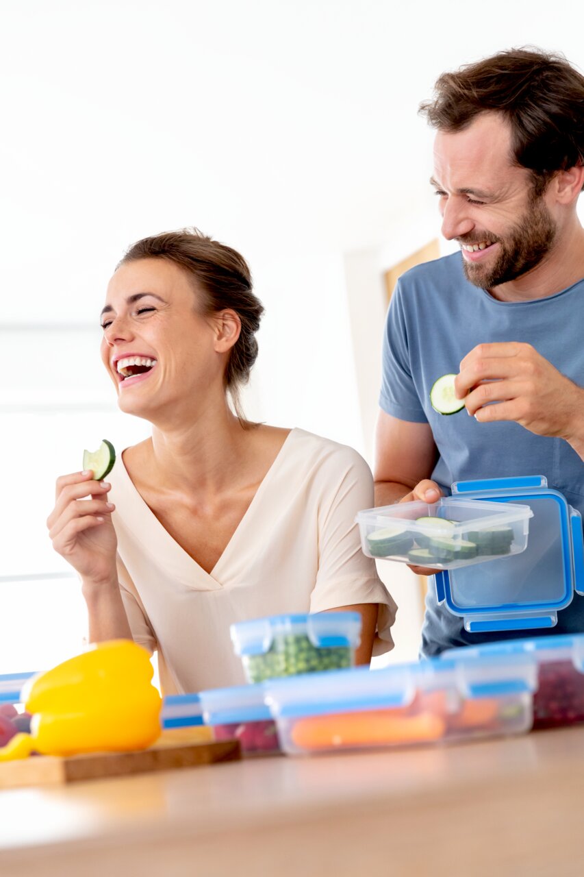 Zwei lachende Personen in einer Küche, die Frischhaltedosen mit geschnittenem Gemüse verwenden. Die Perspektive zeigt die Dosen von vorne.