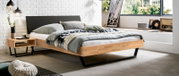 Modernes Schlafzimmer mit gepolstertem Kopfteil, seitliche Perspektive. Das Bett hat ein helles Holzgestell und ist mit grauen und grünen Kissen sowie einer gemusterten Decke dekoriert. Neben dem Bett steht ein Nachttisch mit Dekorationen.