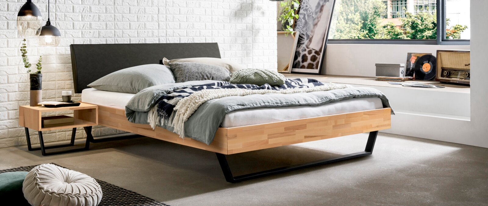 Modernes Schlafzimmer mit gepolstertem Kopfteil, seitliche Perspektive. Das Bett hat ein helles Holzgestell und ist mit grauen und grünen Kissen sowie einer gemusterten Decke dekoriert. Neben dem Bett steht ein Nachttisch mit Dekorationen.