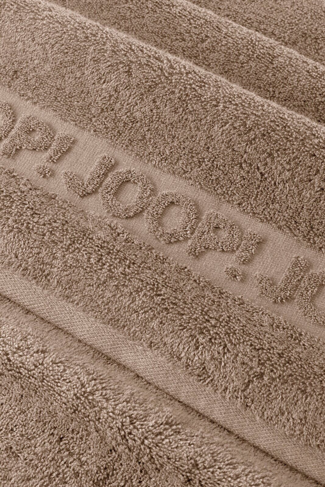 Makro-Nahaufnahme aus schräger Draufsicht eines taupefarbenen Frottee-Handtuchs mit eingewebtem Schriftzug „JOOP!“ und weicher, strukturierter Oberfläche