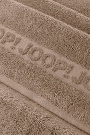 Makro-Nahaufnahme aus schräger Draufsicht eines taupefarbenen Frottee-Handtuchs mit eingewebtem Schriftzug „JOOP!“ und weicher, strukturierter Oberfläche