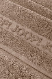 Makro-Nahaufnahme aus schräger Draufsicht eines taupefarbenen Frottee-Handtuchs mit eingewebtem Schriftzug „JOOP!“ und weicher, strukturierter Oberfläche