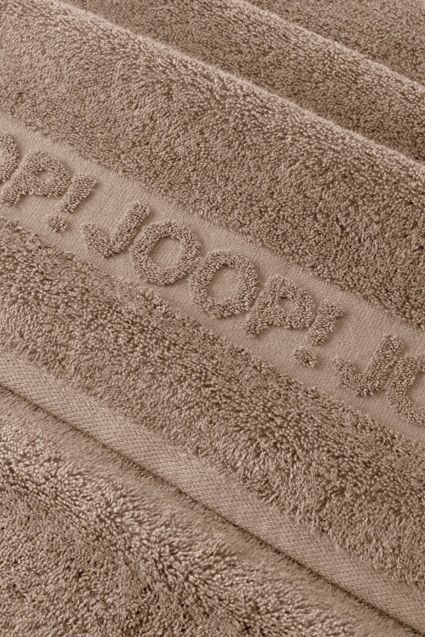 Makro-Nahaufnahme aus schräger Draufsicht eines taupefarbenen Frottee-Handtuchs mit eingewebtem Schriftzug „JOOP!“ und weicher, strukturierter Oberfläche