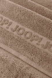 Makro-Nahaufnahme aus schräger Draufsicht eines taupefarbenen Frottee-Handtuchs mit eingewebtem Schriftzug „JOOP!“ und weicher, strukturierter Oberfläche