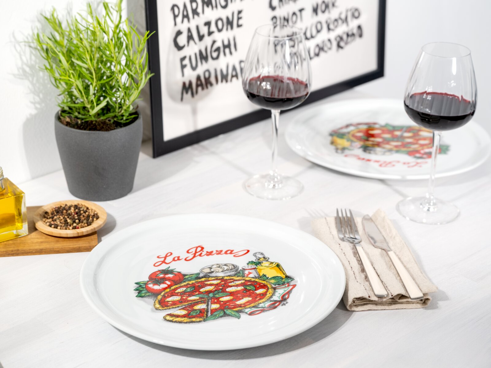 Weißer Keramik-Pizzateller mit farbigem „La Pizza“-Motiv auf gedecktem Tisch, fotografiert aus leicht schräger Draufsicht (3/4-Perspektive) mit Weingläsern im Hintergrund.