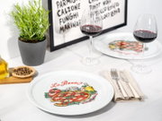 Weißer Keramik-Pizzateller mit farbigem „La Pizza“-Motiv auf gedecktem Tisch, fotografiert aus leicht schräger Draufsicht (3/4-Perspektive) mit Weingläsern im Hintergrund.