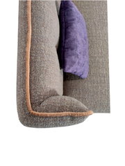 Detailansicht eines grauen, strukturierten Sofas mit beigefarbener Kederpaspel und violettem Kissen, seitliche Perspektive