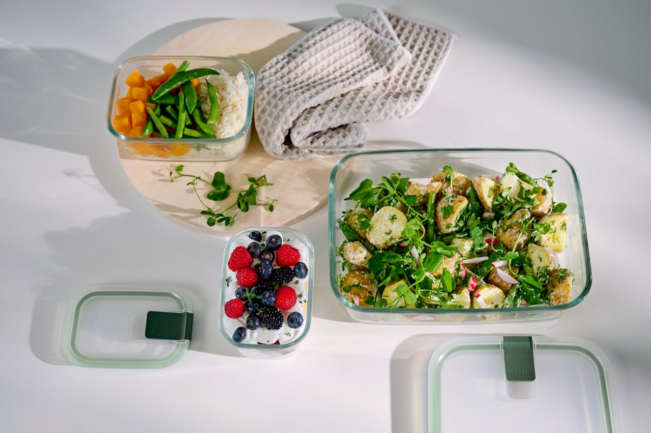 Vogelperspektive auf Glas-Frischhaltedosen mit verschiedenen Speisen, darunter Salat, Obst und Gemüse, auf einem weißen Hintergrund.