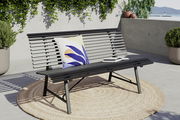 Moderne Gartenbank aus Metall mit Kissen und Buch, seitliche Perspektive, auf einer Terrasse mit Pflanzen im Hintergrund.