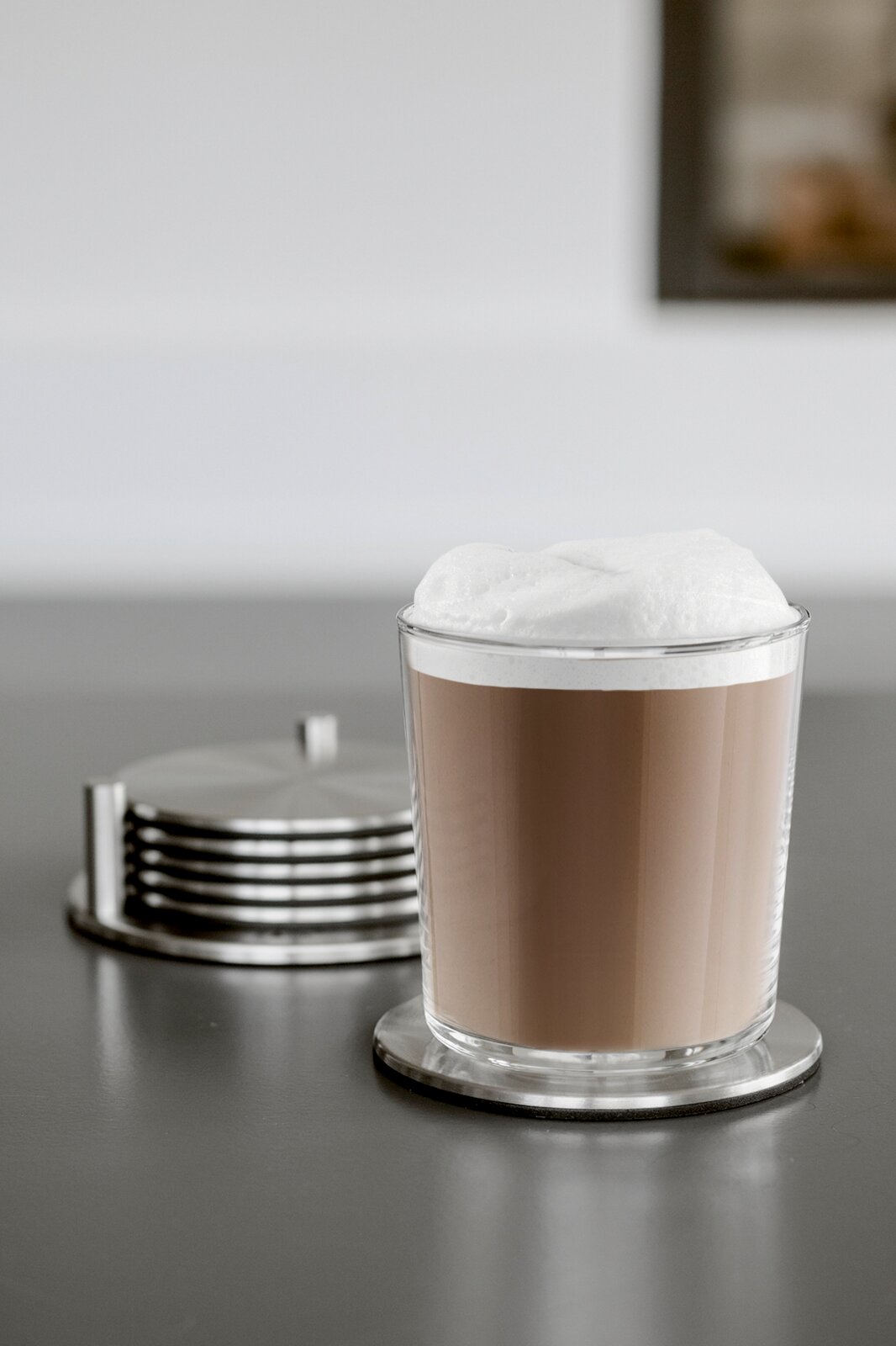 Transparenter Glasbecher mit Latte Macchiato und cremigem Milchschaum auf rundem Untersetzer, seitliche Frontansicht auf einer grauen Tischfläche; im Hintergrund ein Stapel Metall-Untersetzer unscharf.