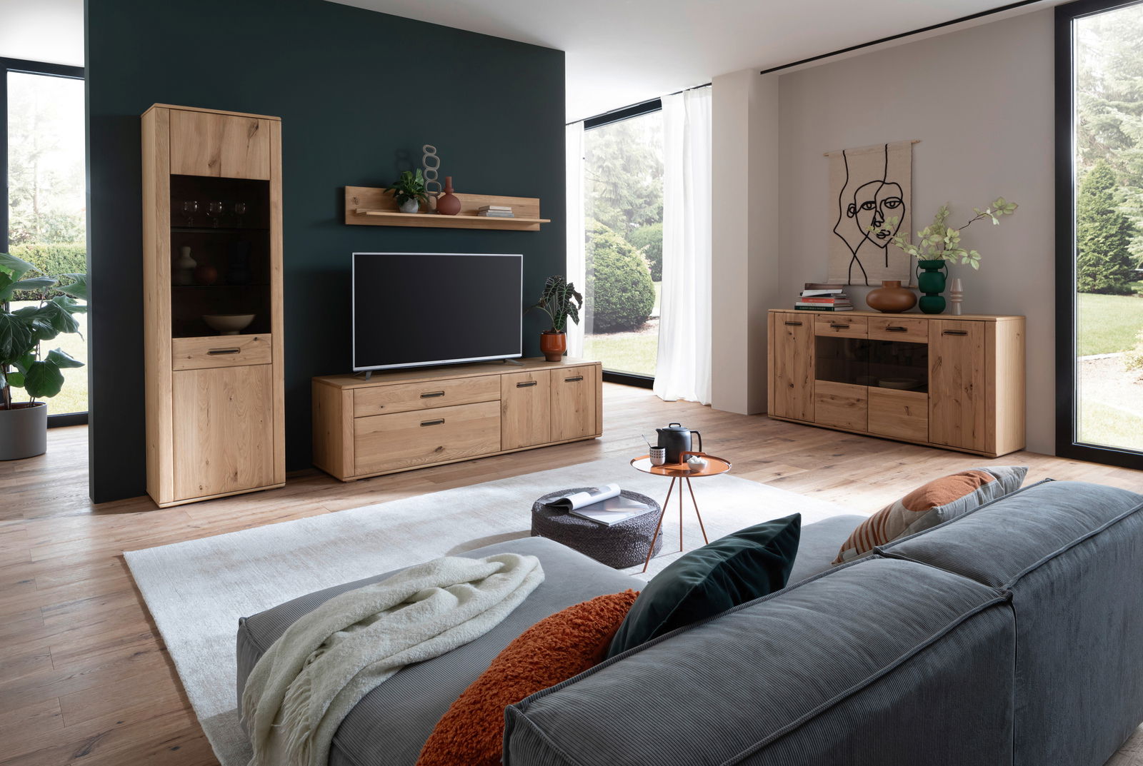 Moderne Wohnzimmeransicht mit Cantus Wandkombination ASINARA in Holzoptik, bestehend aus einem hohen Schrank, einem TV-Board und einem Sideboard, mit Blick auf große Fenster und stilvolle Dekoration.