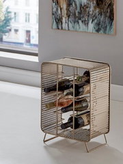 Metallischer Weinflaschenhalter in goldfarbenem Lochblech-Design mit mehreren horizontalen Ablagen, seitlich auf einem weißen Sideboard neben einem Fenster platziert, fotografiert in leichter Seitenansicht.