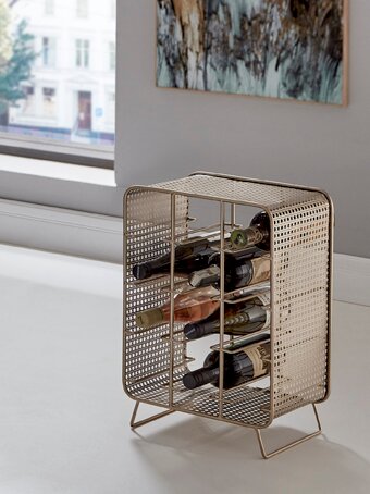 Metallischer Weinflaschenhalter in goldfarbenem Lochblech-Design mit mehreren horizontalen Ablagen, seitlich auf einem weißen Sideboard neben einem Fenster platziert, fotografiert in leichter Seitenansicht.