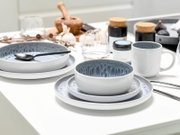 Kombiservice 16-tlg. Stone auf einem Tisch, bestehend aus Tellern, Schalen und einer Tasse in modernem Design mit grauem und weißem Farbverlauf, seitliche Perspektive.