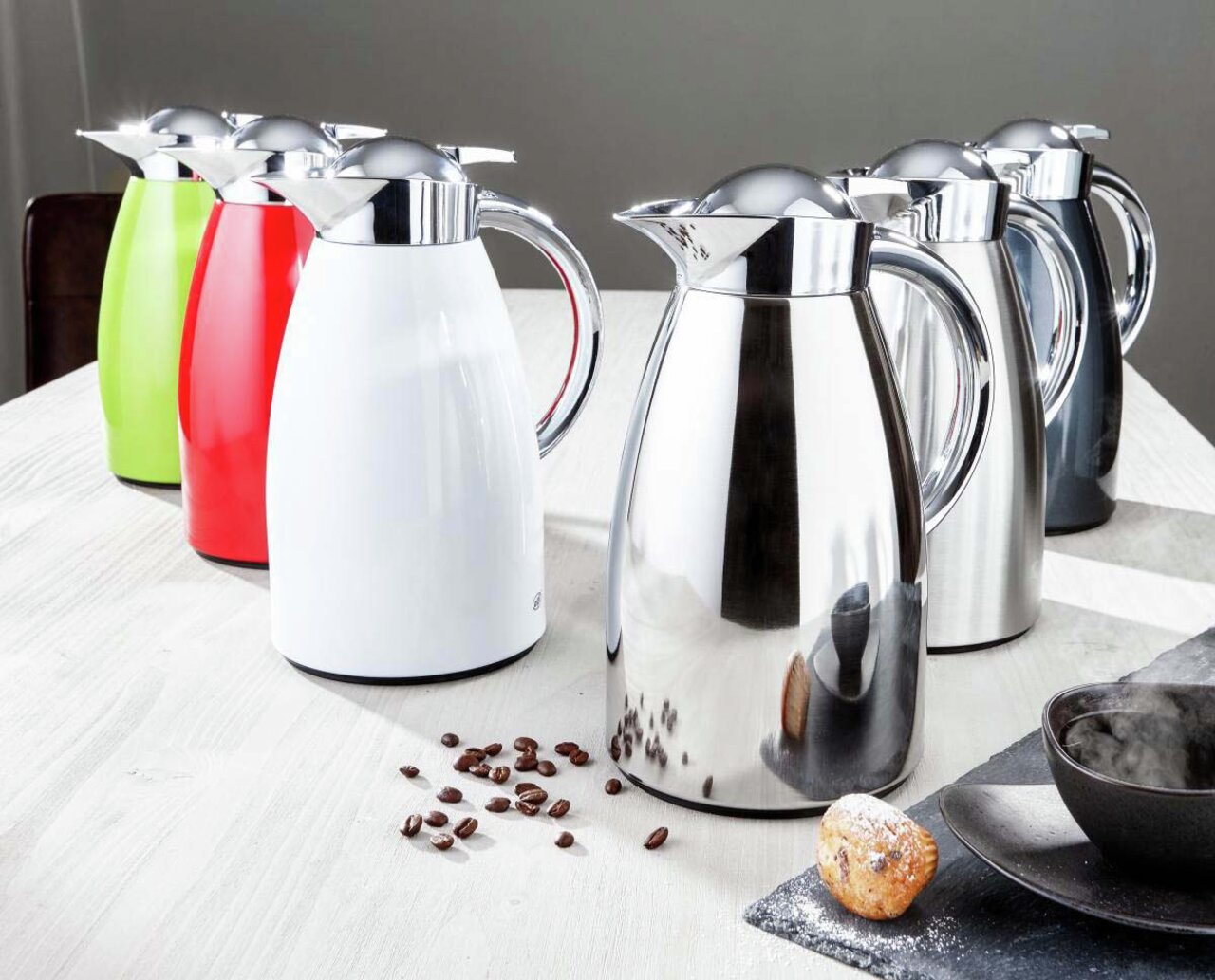 Mehrere Isolierkannen Signo aus Edelstahl in verschiedenen Farben auf einem Tisch, seitliche Perspektive.