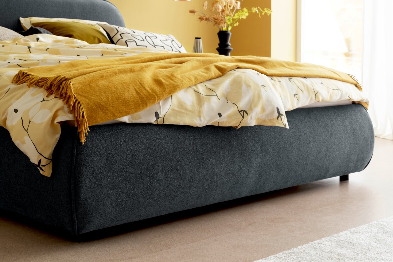 Seitliche Perspektive eines modernen Polsterbetts mit dunkelgrauem Stoffbezug, gelber Decke und gemustertem Bettzeug in einem hellen Raum.