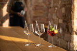 Drei elegante Gin Tonic-Gläser auf einem Holztisch, eines gefüllt mit einem roten Getränk und Minzblättern, im Hintergrund eine unscharfe Person. Seitliche Perspektive.