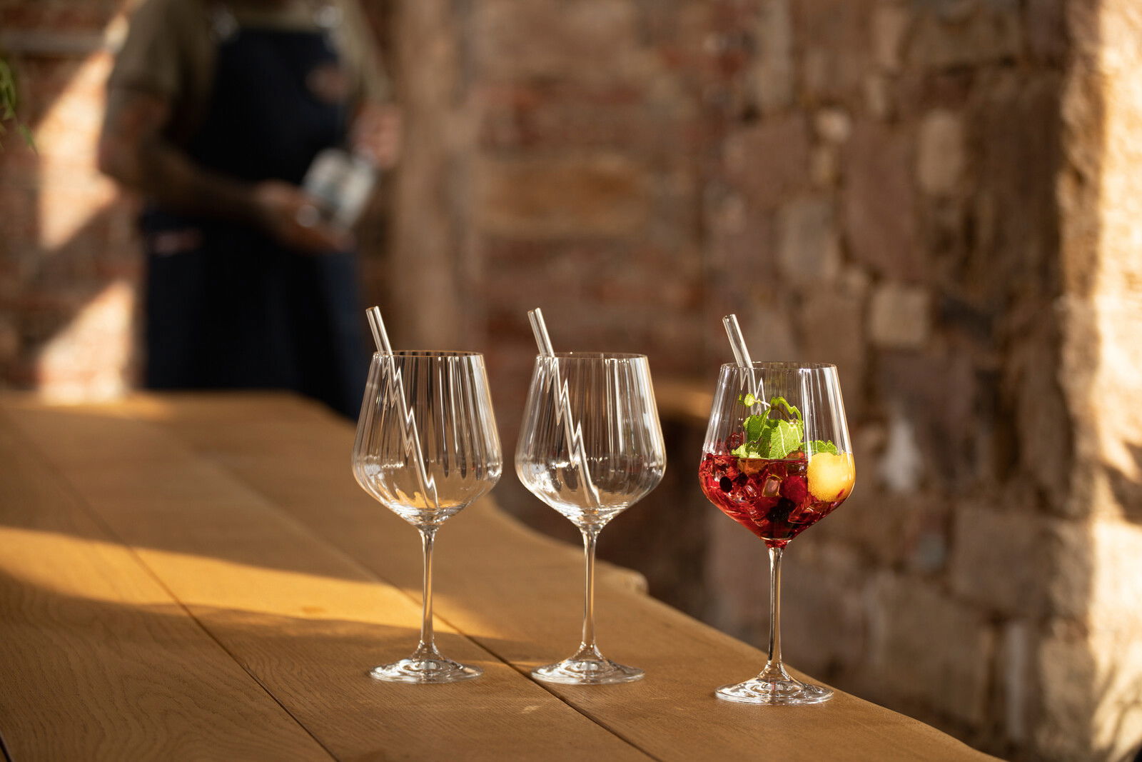 Drei elegante Gin Tonic-Gläser auf einem Holztisch, eines gefüllt mit einem roten Getränk und Minzblättern, im Hintergrund eine unscharfe Person. Seitliche Perspektive.