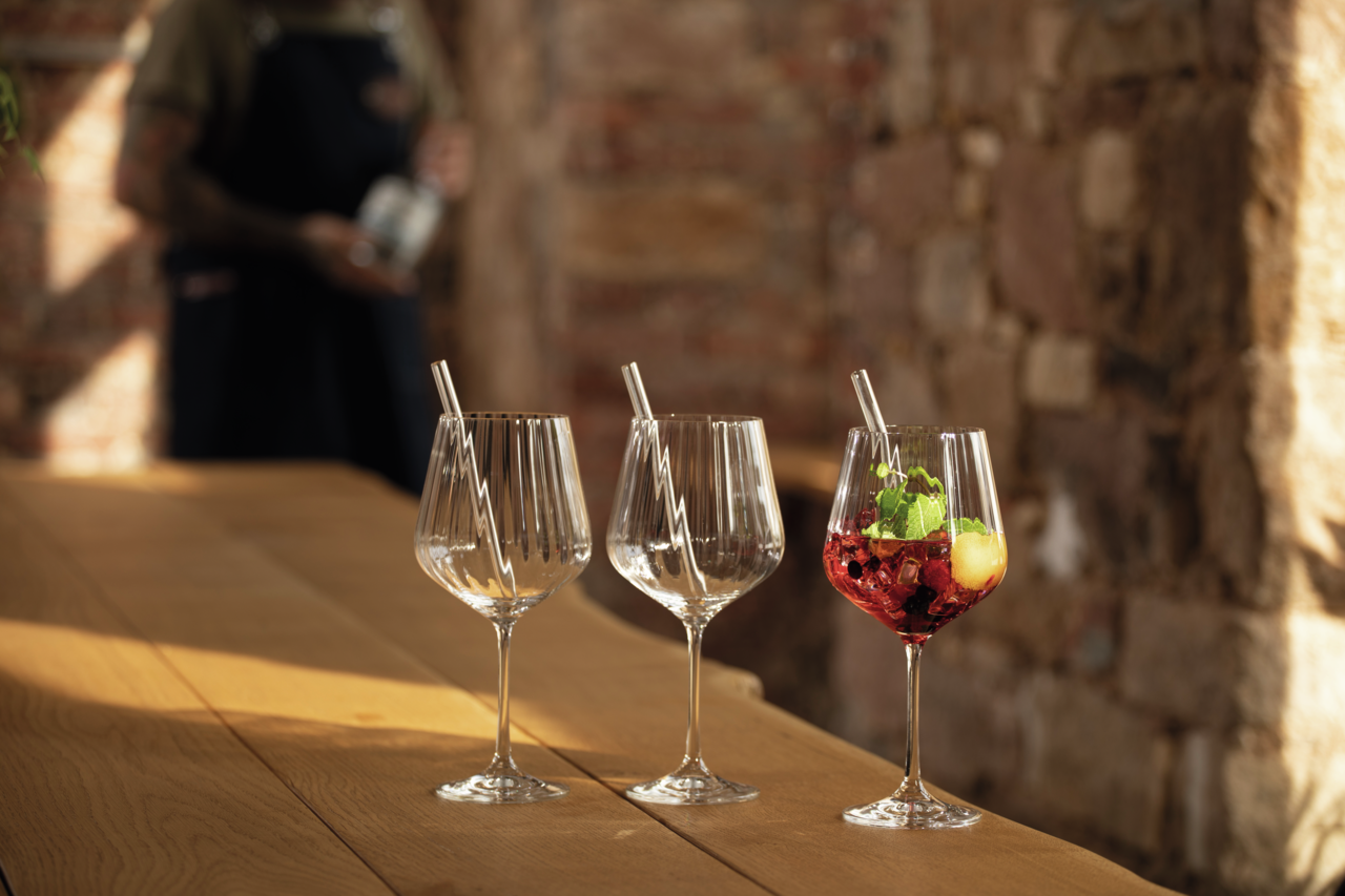 Drei elegante Gin Tonic-Gläser auf einem Holztisch, eines gefüllt mit einem roten Getränk und Minzblättern, im Hintergrund eine unscharfe Person. Seitliche Perspektive.