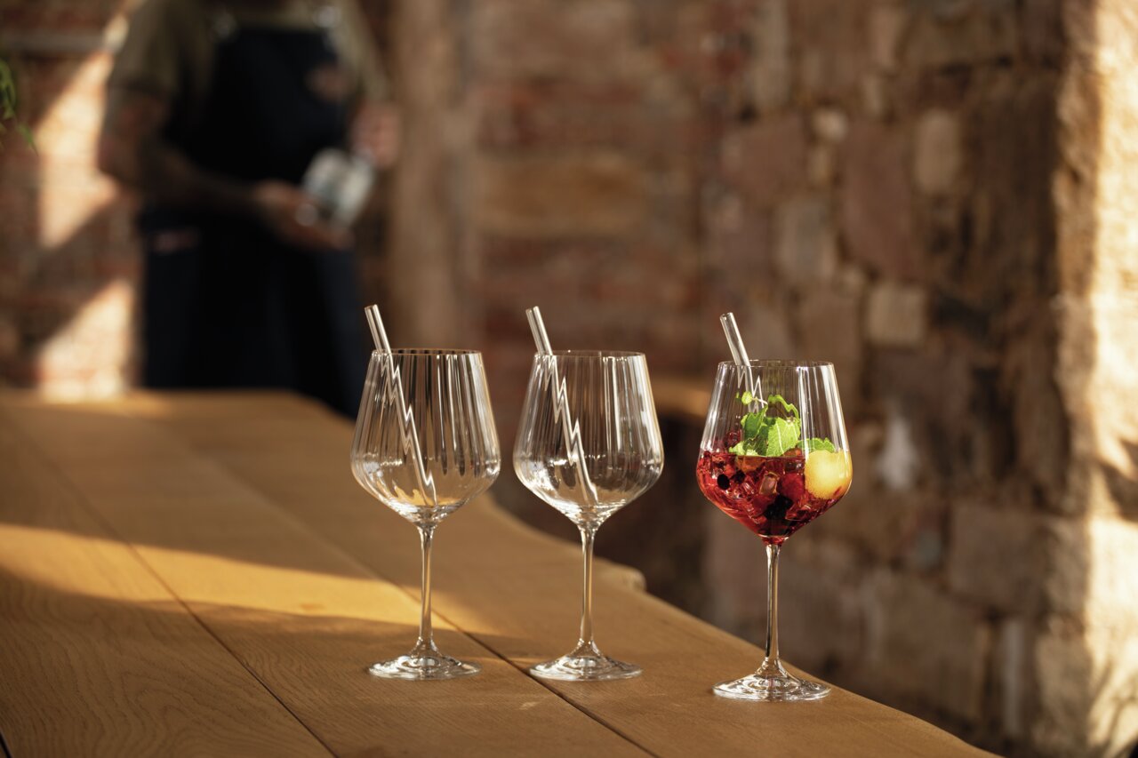 Drei elegante Gin Tonic-Gläser auf einem Holztisch, eines gefüllt mit einem roten Getränk und Minzblättern, im Hintergrund eine unscharfe Person. Seitliche Perspektive.