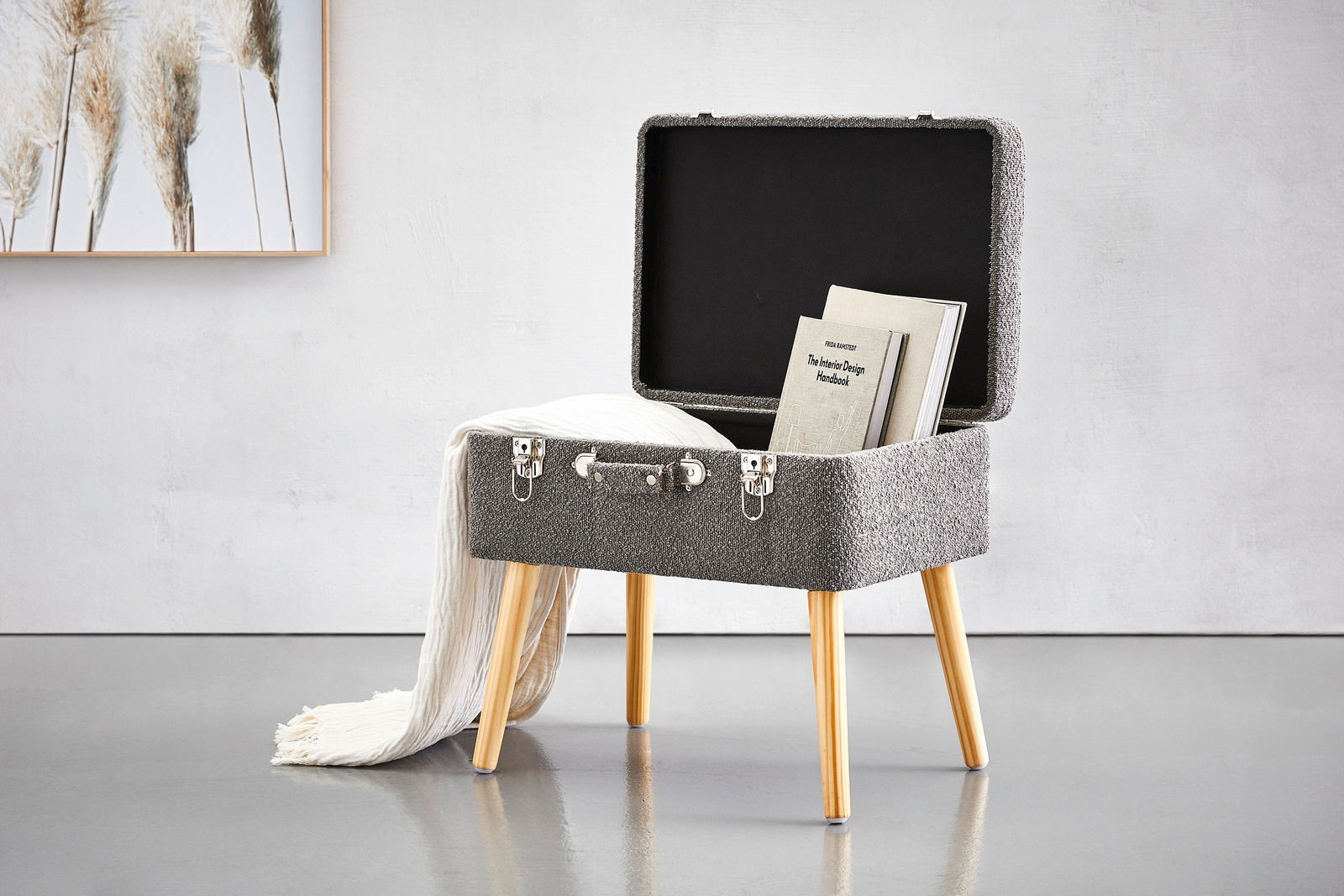 Kofferhocker mit geöffnetem Deckel, aus grauem Stoff mit Holzbeinen, von vorne betrachtet. Im Inneren befinden sich Bücher und eine weiße Decke hängt über der Seite.