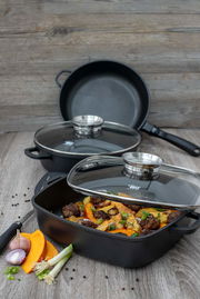 Alucast Servierpfanne rund 28 cm mit Glasdeckel, gefüllt mit gebratenem Gemüse und Fleisch, auf einem Holztisch aus der Seitenperspektive fotografiert.