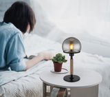 Moderne Tischleuchte HAKU mit rauchgrauem Glasschirm und Holzfuß, auf einem Tisch neben einer Pflanze, seitliche Perspektive.