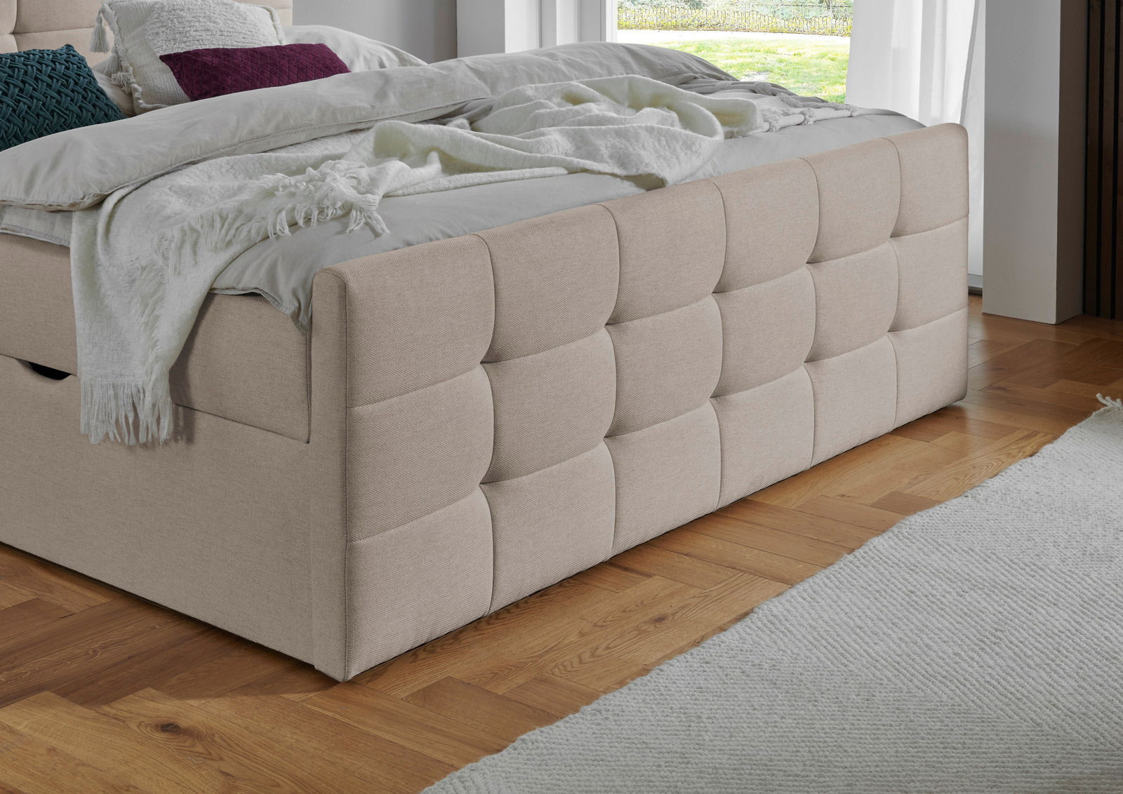 Beige Boxbett mit gestepptem Kopfteil, seitlicher Blick auf das Bett in einem modernen Schlafzimmer mit Holzboden.