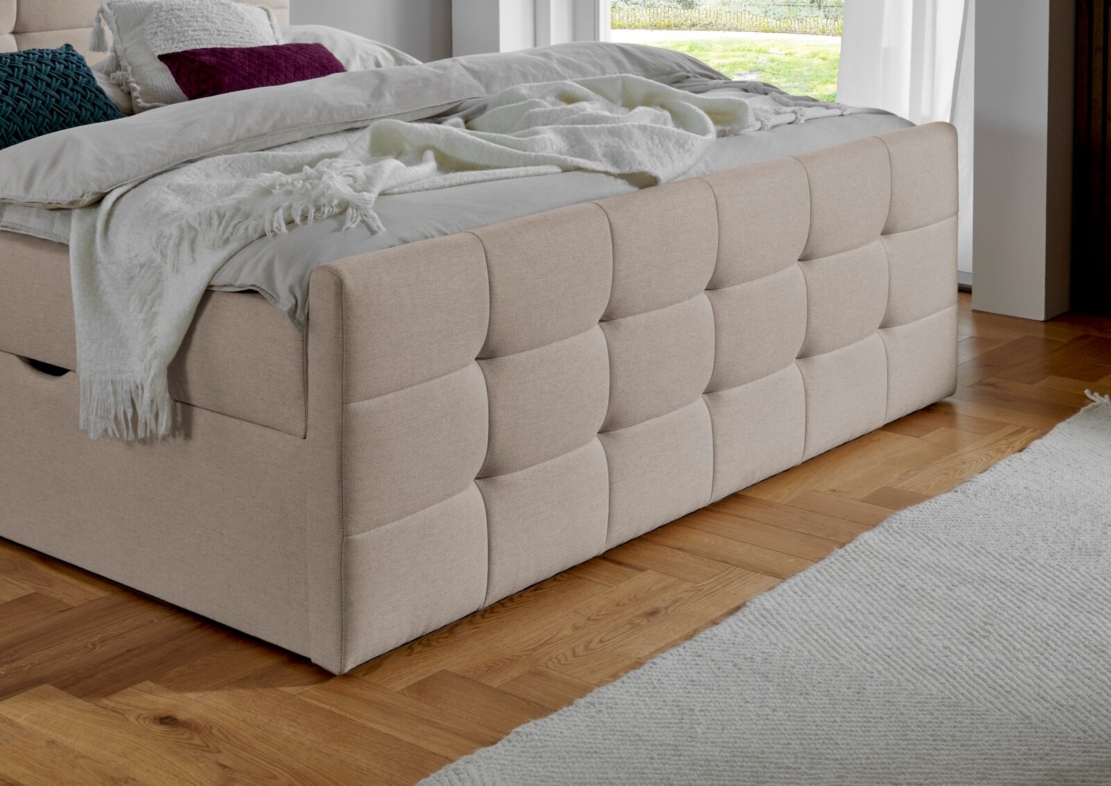 Beige boxspring met doorgestikt hoofdeinde, zijaanzicht van het bed in een moderne slaapkamer met houten vloer.