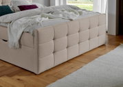 Beige boxspring met doorgestikt hoofdeinde, zijaanzicht van het bed in een moderne slaapkamer met houten vloer.
