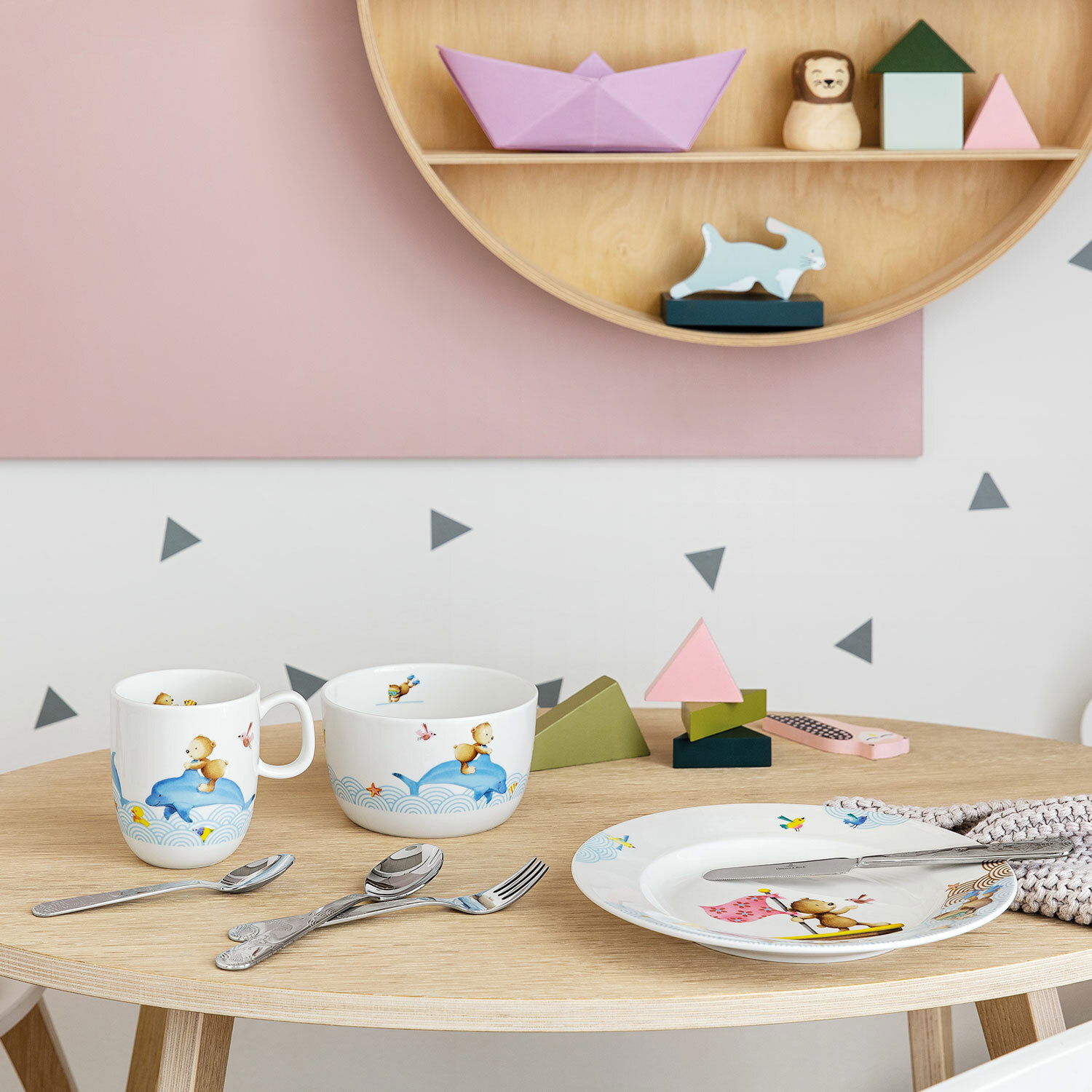 Kinderbesteck-Set mit bunten Tiermotiven, bestehend aus Tasse, Schüssel, Teller und Besteck, auf einem Holztisch in einer verspielten Umgebung präsentiert. Perspektive: Frontalansicht.