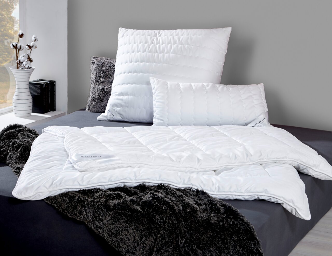 Faserdecke Duo ANTIGUA von billerbeck auf einem Bett, umgeben von Kissen und einer flauschigen Decke, seitliche Perspektive