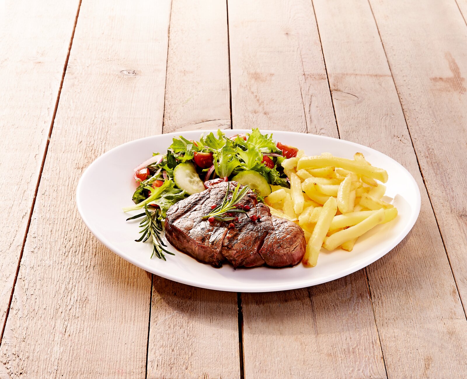 Weißer Teller mit gebratenem Steak mit Rosmarin, Pommes frites und gemischtem Salat auf Holztisch, schräg von oben fotografiert.