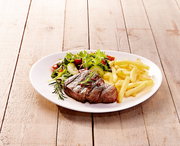 Weißer Teller mit gebratenem Steak mit Rosmarin, Pommes frites und gemischtem Salat auf Holztisch, schräg von oben fotografiert.