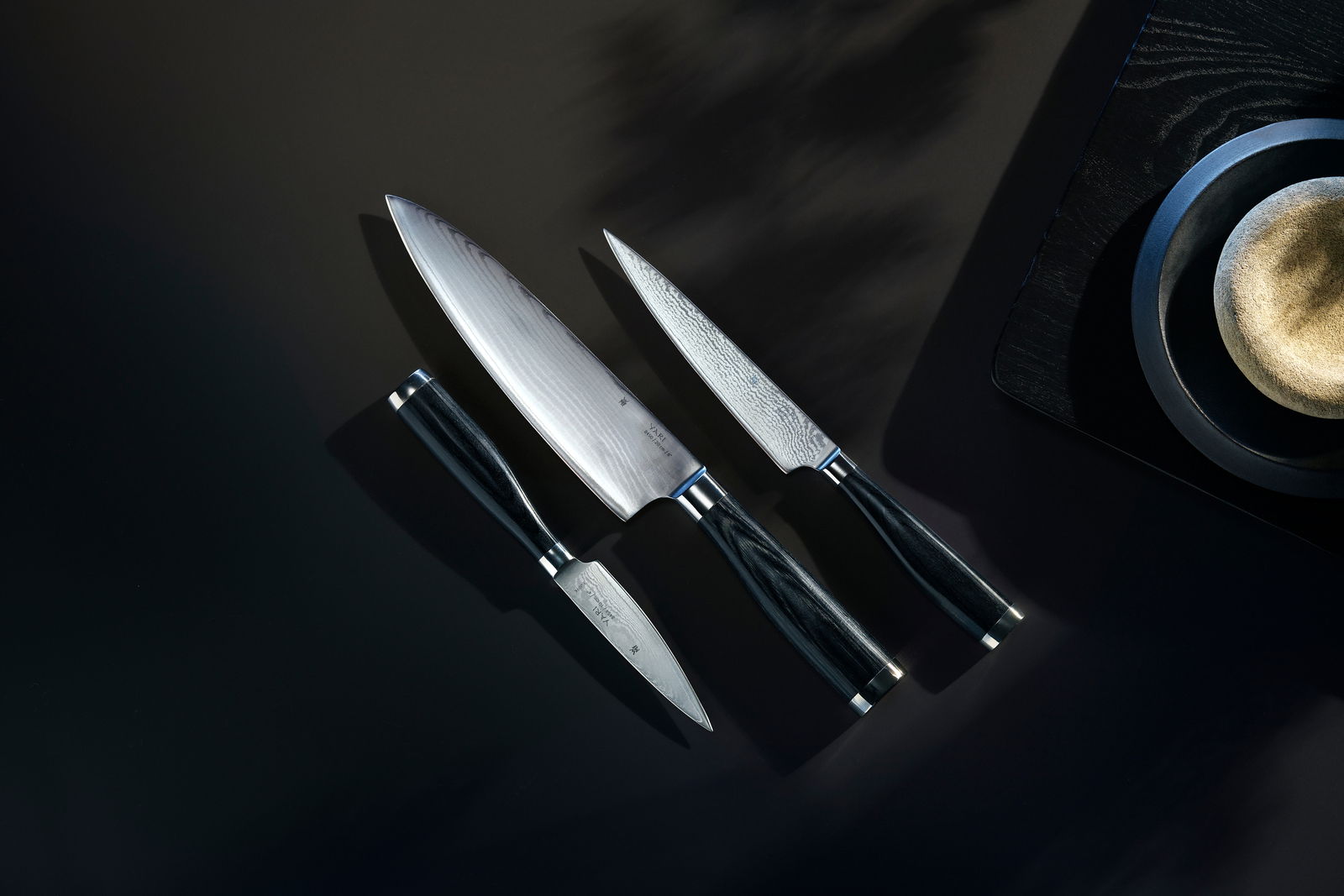 Dreiteiliges Messerset Yari aus der Vogelperspektive, bestehend aus einem großen Kochmesser, einem mittleren Allzweckmesser und einem kleinen Schälmesser, auf dunklem Hintergrund.