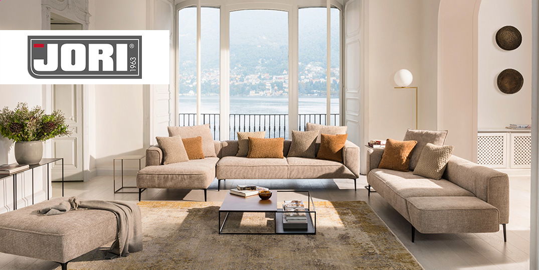 Wohnzimmer mit einer stilvollen beigen Sofagarnitur, einem Couchtisch aus Metall, großen Fenstern und einem Teppich in unterschiedlichen Beigetönen.