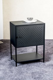 Schwarzer Metall-Nachttisch/Beistellschrank mit perforierter Fronttür, rundem Griff und unterer Ablage, frontal in leichter Schrägansicht von oben fotografiert; oben steht ein Teller mit kleiner Schale.