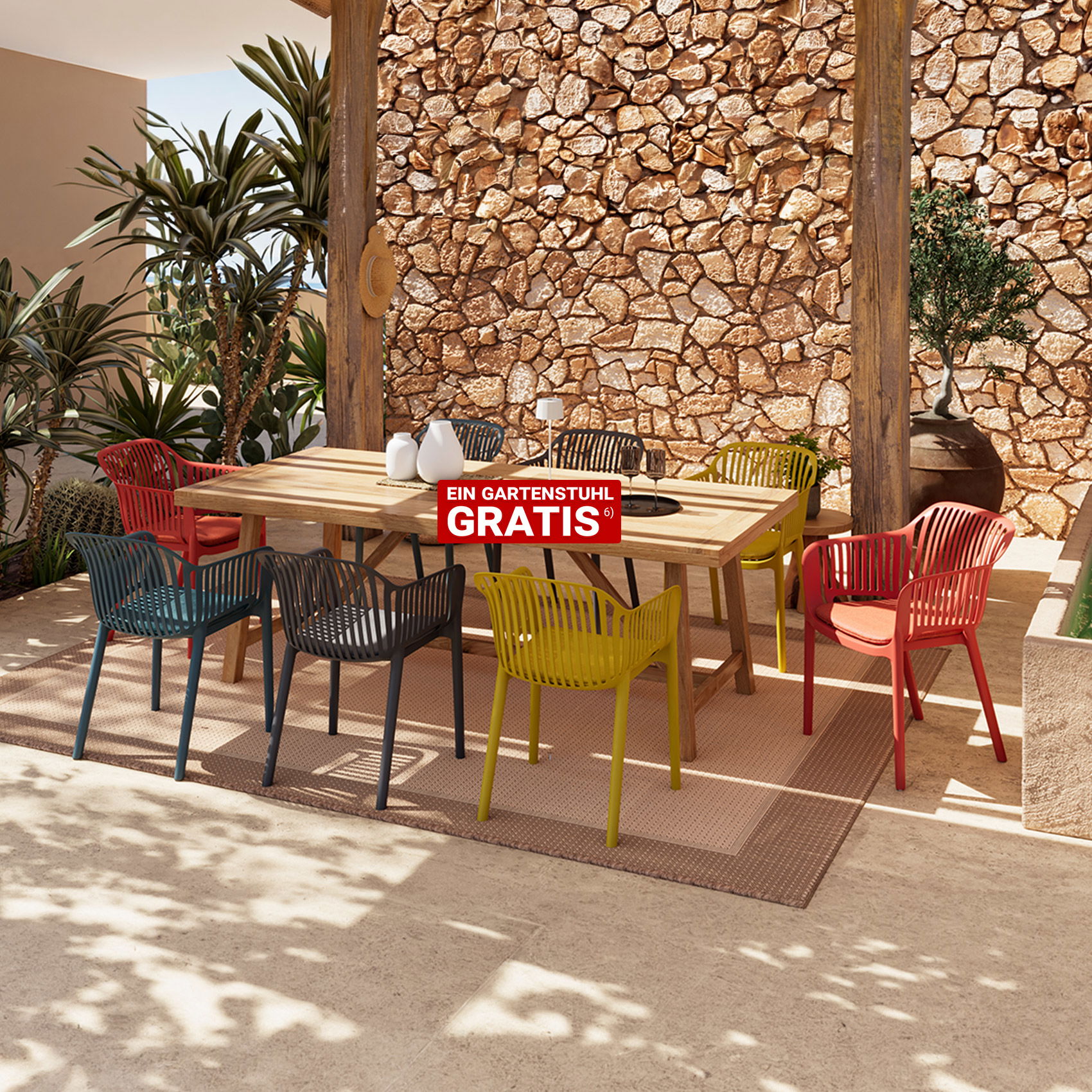 Großer rechteckiger Holztisch mit mehreren bunten Kunststoffstühlen auf einer Terrasse vor einer Steinwand und Pflanzen