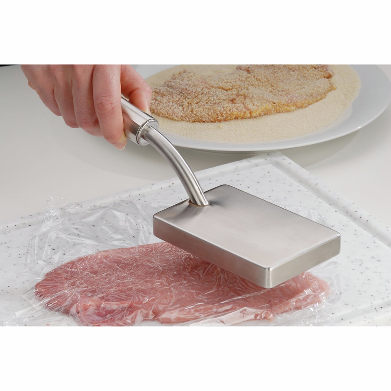 Fleischklopfer PROFI PLUS aus Edelstahl, in der Hand gehalten, von oben auf ein Stück Fleisch auf einem Schneidebrett gedrückt.