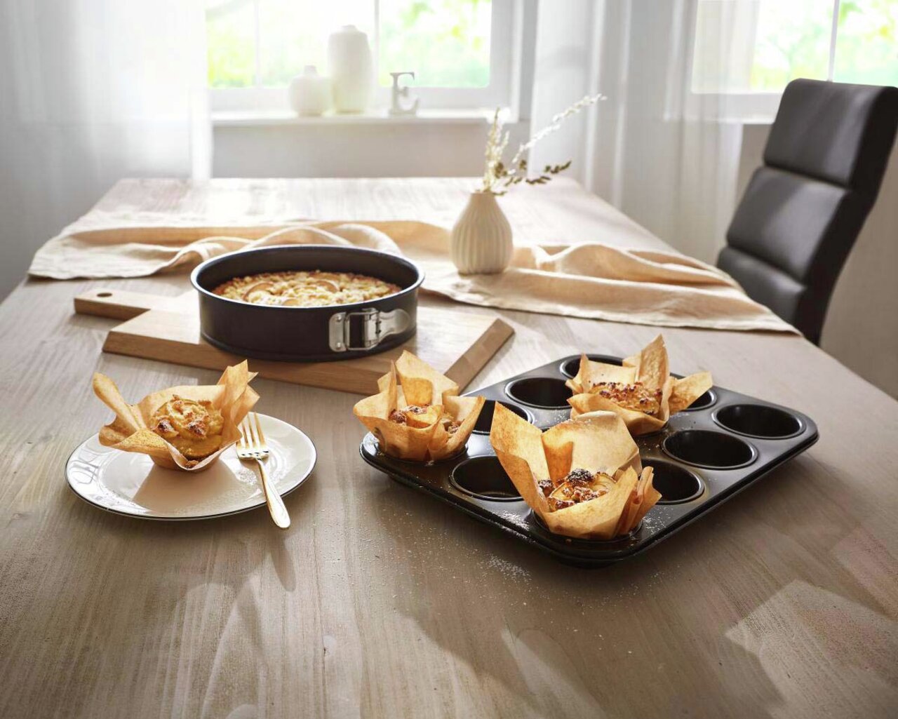 Springform 26 cm auf einem Tisch mit gebackenem Kuchen, umgeben von Muffins in einer Muffinform, seitliche Perspektive