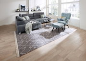 Woonkamer met een grijze hoekbank en een lichtblauwe fauteuil met bijpassend voetenbankje, gezien vanaf de voorkant. Moderne inrichting met tapijt en bijzettafels.