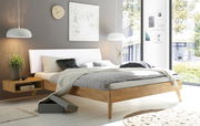 Modernes Schlafzimmer mit gepolstertem Kopfteil, seitliche Perspektive. Helles Holzgestell, graue Bettwäsche und dekorative Kissen. Große Fenster mit Blick ins Grüne.