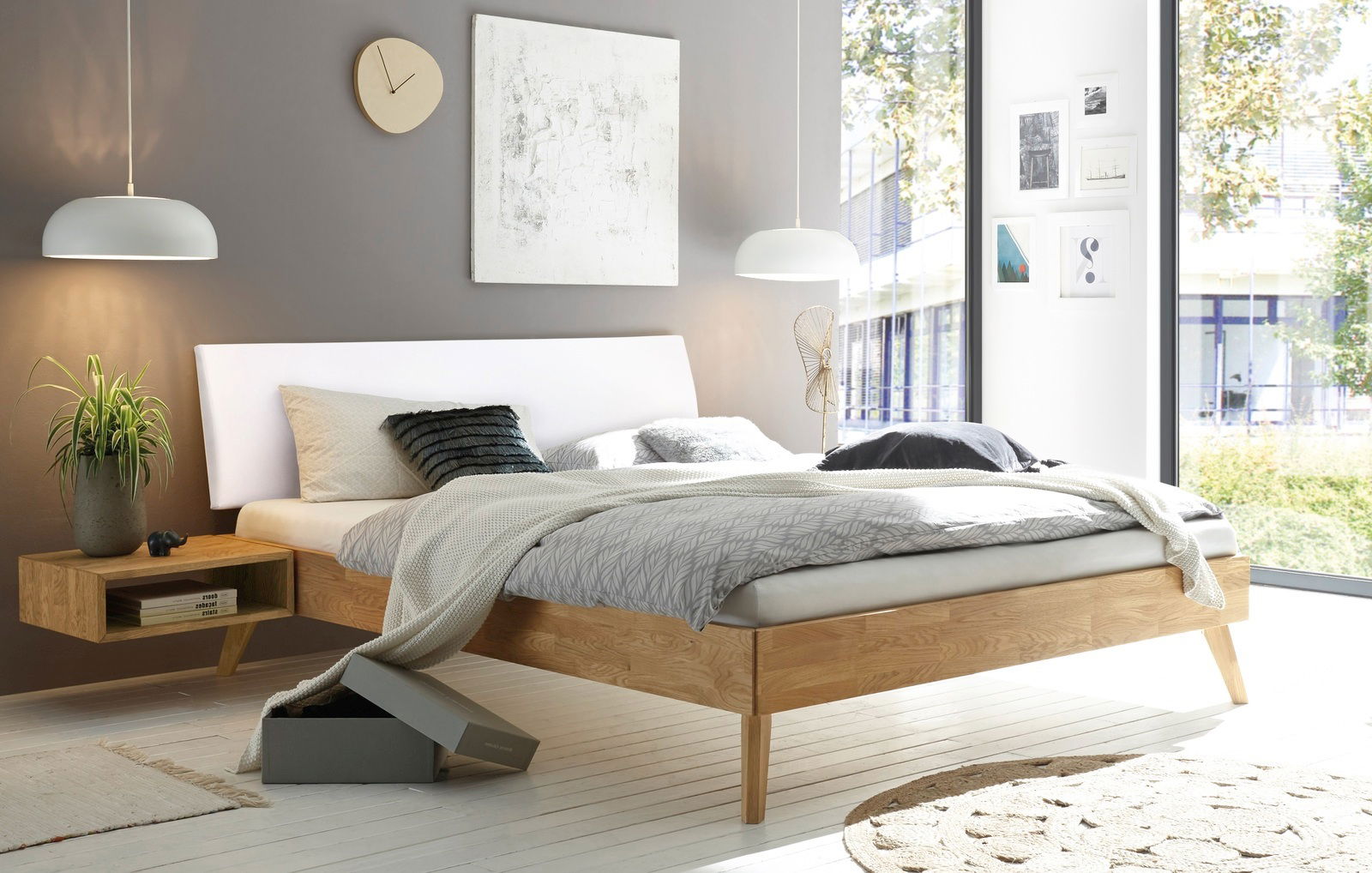 Modernes Schlafzimmer mit gepolstertem Kopfteil, seitliche Perspektive. Helles Holzgestell, graue Bettwäsche und dekorative Kissen. Große Fenster mit Blick ins Grüne.