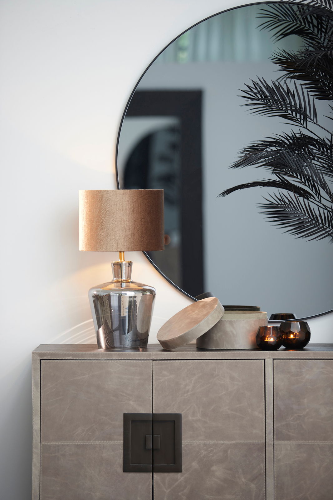 Moderner Lampenschirm in Zylinderform auf einem eleganten Glastisch, mit einem großen runden Spiegel im Hintergrund. Perspektive von vorne.