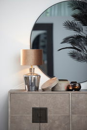 Moderner Lampenschirm in Zylinderform auf einem eleganten Glastisch, mit einem großen runden Spiegel im Hintergrund. Perspektive von vorne.