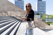 Beige-weiß gestreifte Tragetasche mit kurzen Henkeln, von einer Frau im urbanen Außenbereich getragen; seitliche Dreiviertelansicht der Tasche.
